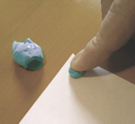 A finger presses a small blob of adhesive putty onto the corner of a sheet of paper. A larger blob sits on the table.